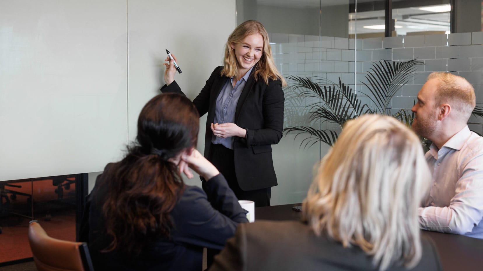 Kontorlandskap med fire personer i et møterom. En smilende dame i forretningsklær holder en penn og skal tegne på tavlen.
