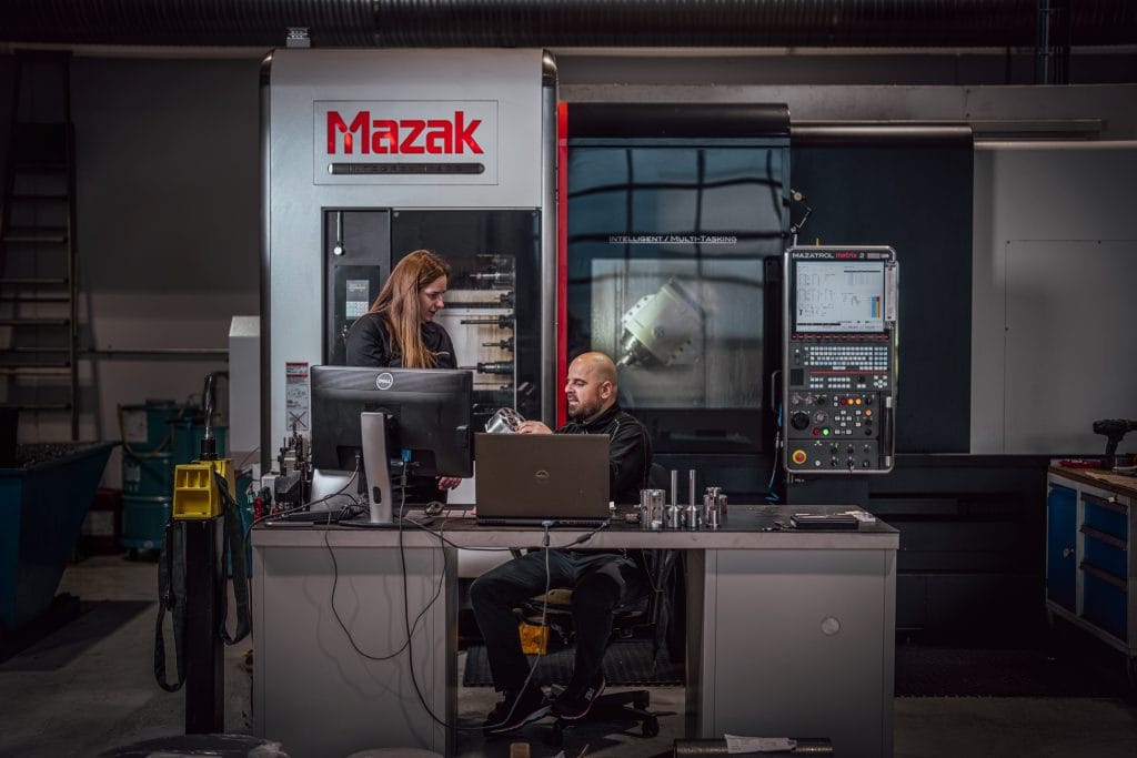 Bilde tatt i en maskineringsbedrift eller mekanisk verksted, inne i en produksjonshall. To mennesker i arbeidsklær står foran en CNC-maskin og bak en pc. De har en diskusjon.