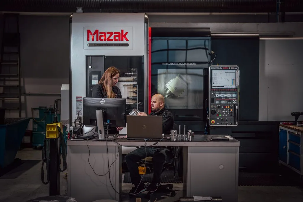 To mennesker i arbeidsklær står foran en mazak-maskin og bak en pc. De har en diskusjon.